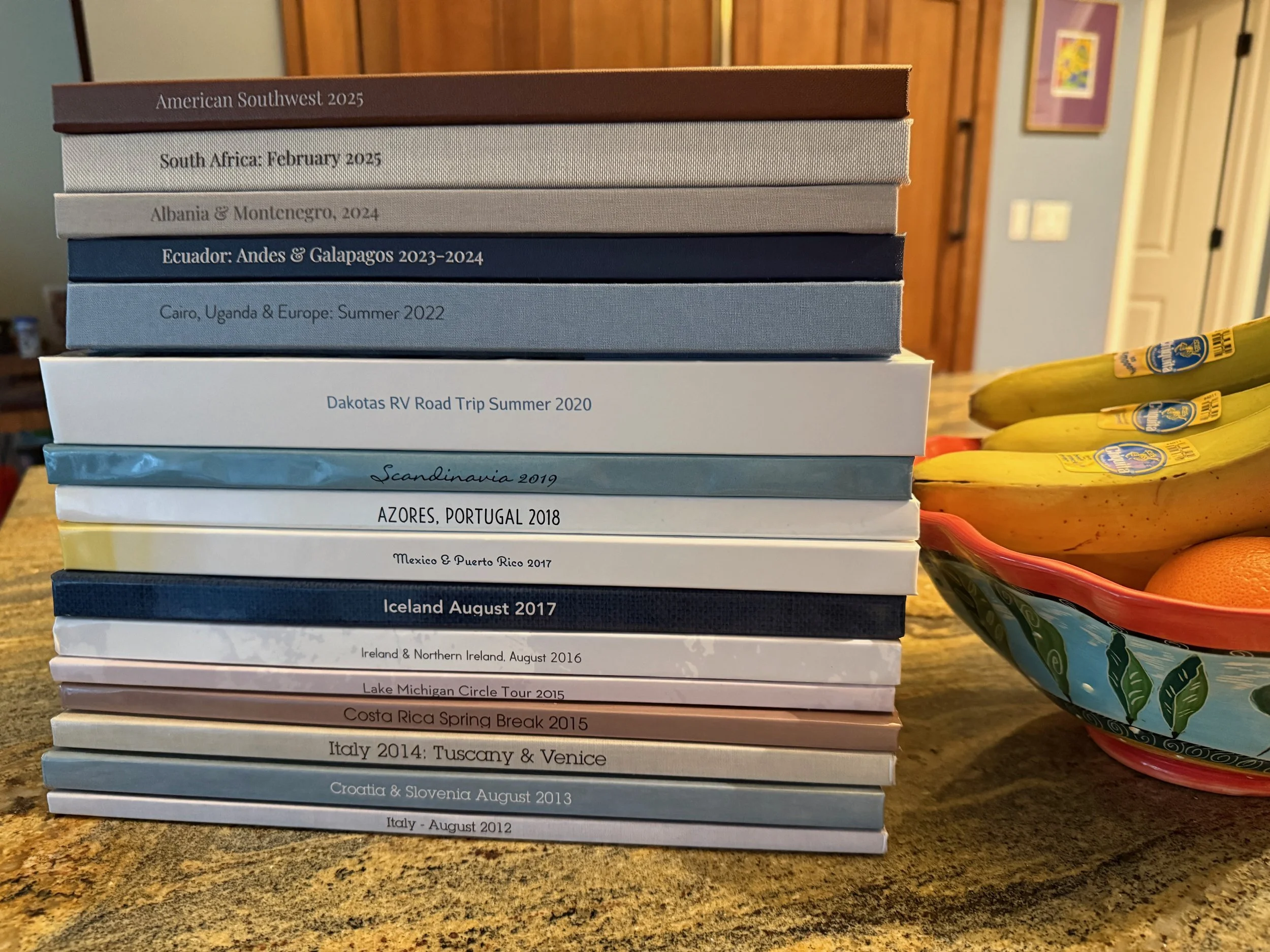stack of travel photo books next to a bowl of fruit. each book titled by date and destination