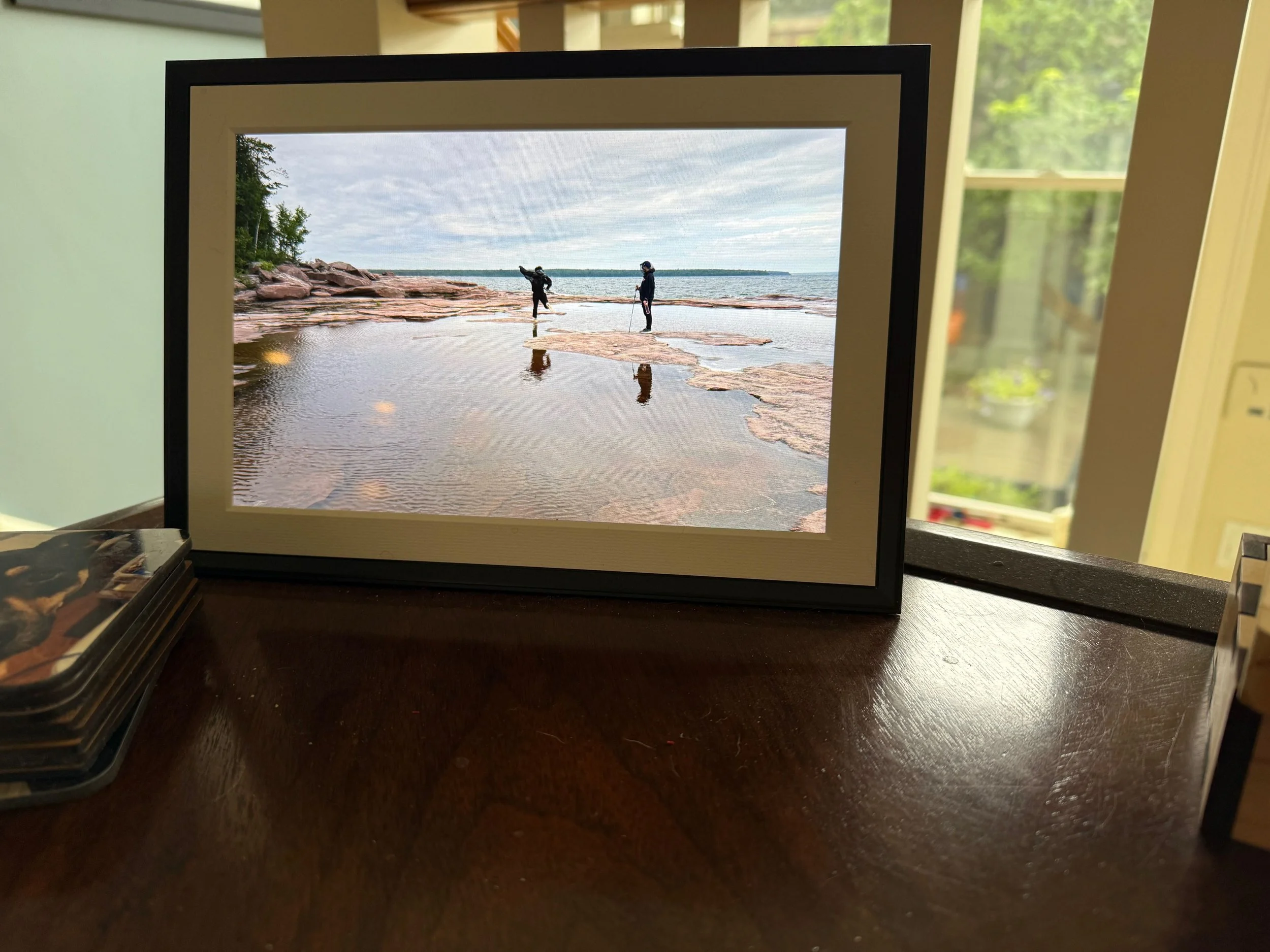 white digital photo frame with black outline showing photo of 2 kids jumping on rocks on a lake. on a brown table.
