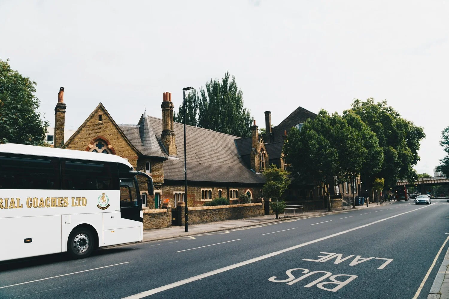 Street view featuring a large white bus labeled 'IMPERIAL COACHES LTD' parked on the side, historic brick buildings with chimneys, a bus lane marked on the road, and trees lining the sidewalk.