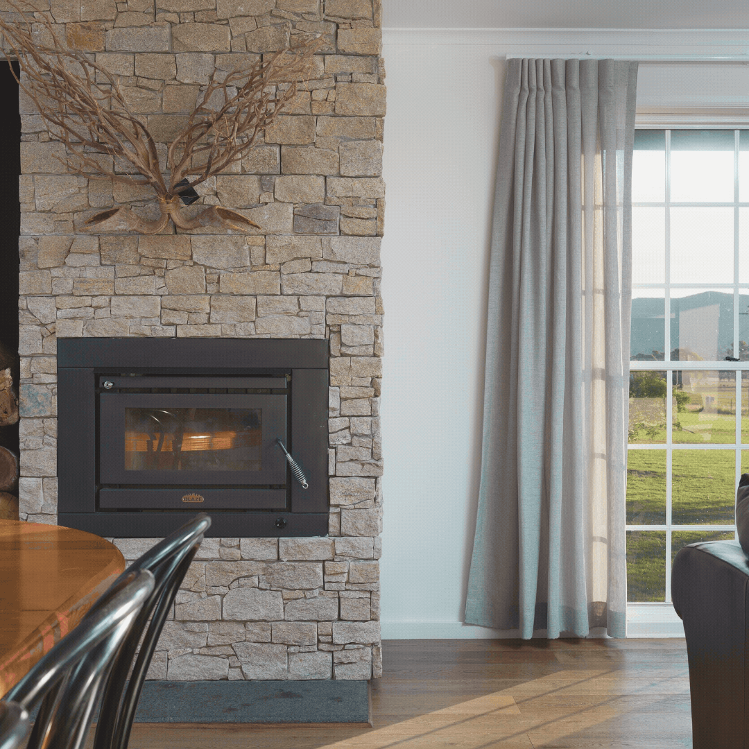 Interior of a living room with a stone fireplace, a mounted animal antler decoration above the fireplace, light gray curtains, a large window showing a scenic outdoor view, and part of a wooden dining table with black chairs.