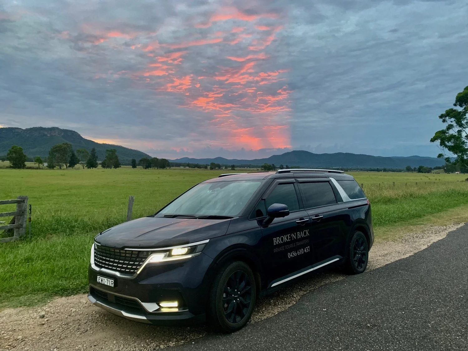 Black SUV parked on the side of a rural road overlooking green fields and distant hills at sunset with pink and blue sky.