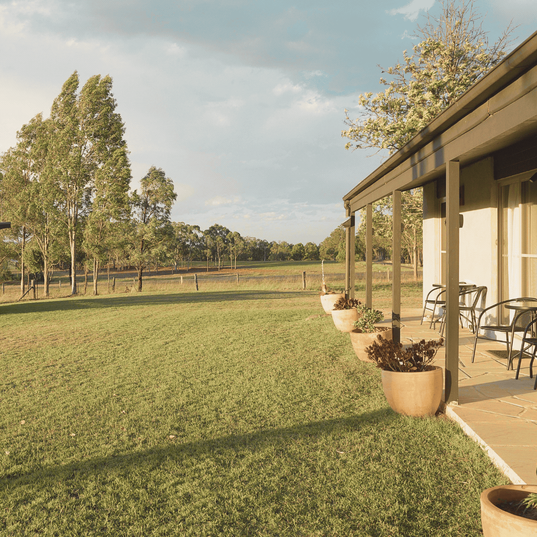 A house with a porch and outdoor chairs, overlooking a grassy field with trees in the distance under a partly cloudy sky.