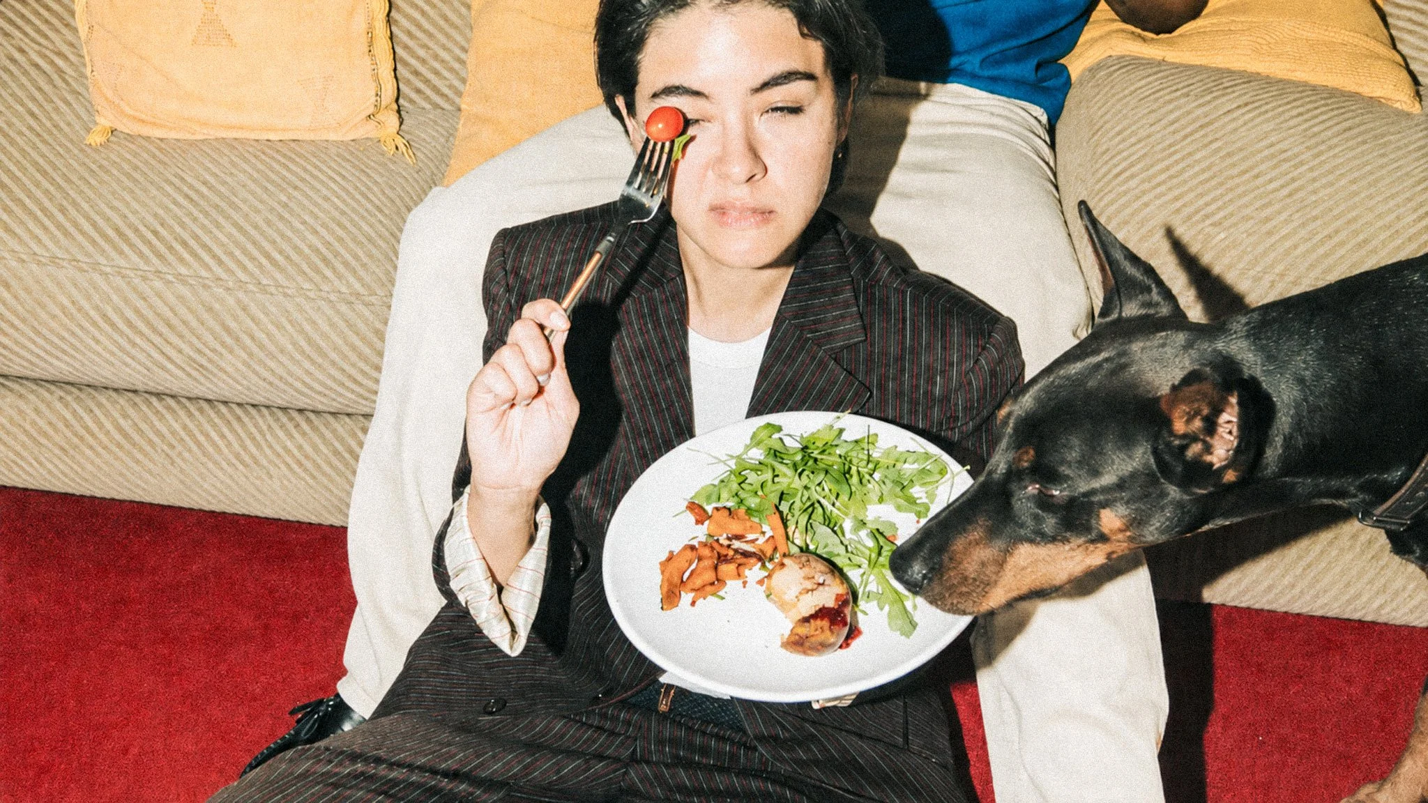 A woman sitting on a red carpeted floor, holding a plate with chicken, a green salad, and some sweet potatoes. She is holding a fork with a cherry tomato in front of her eye, while a Doberman is about to steal her chicken.