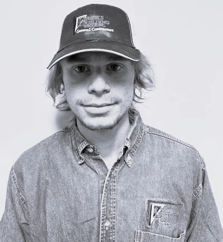 Young man wearing a cap and denim shirt, smiling slightly, against a plain background.
