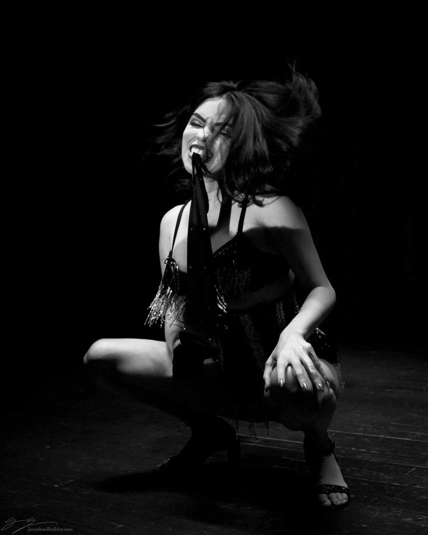 A woman with long hair is performing on stage, squatting with her mouth open, holding a piece of cloth with her teeth, against a dark background.