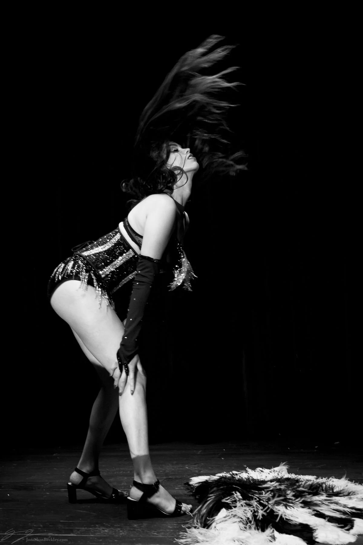 A woman dancing on stage with her hair flying, wearing a sequined top, shorts, and high heels, with feathers on the floor in front of her.