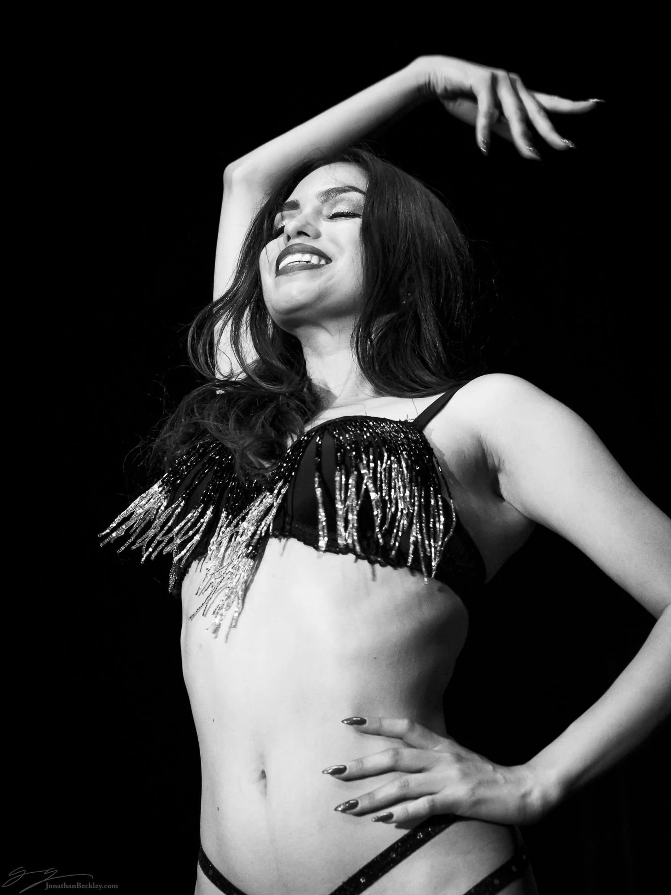 A woman smiling with eyes closed, wearing a fringed, sequined top, and striking a confident pose with one hand on her hip and the other raised above her head.