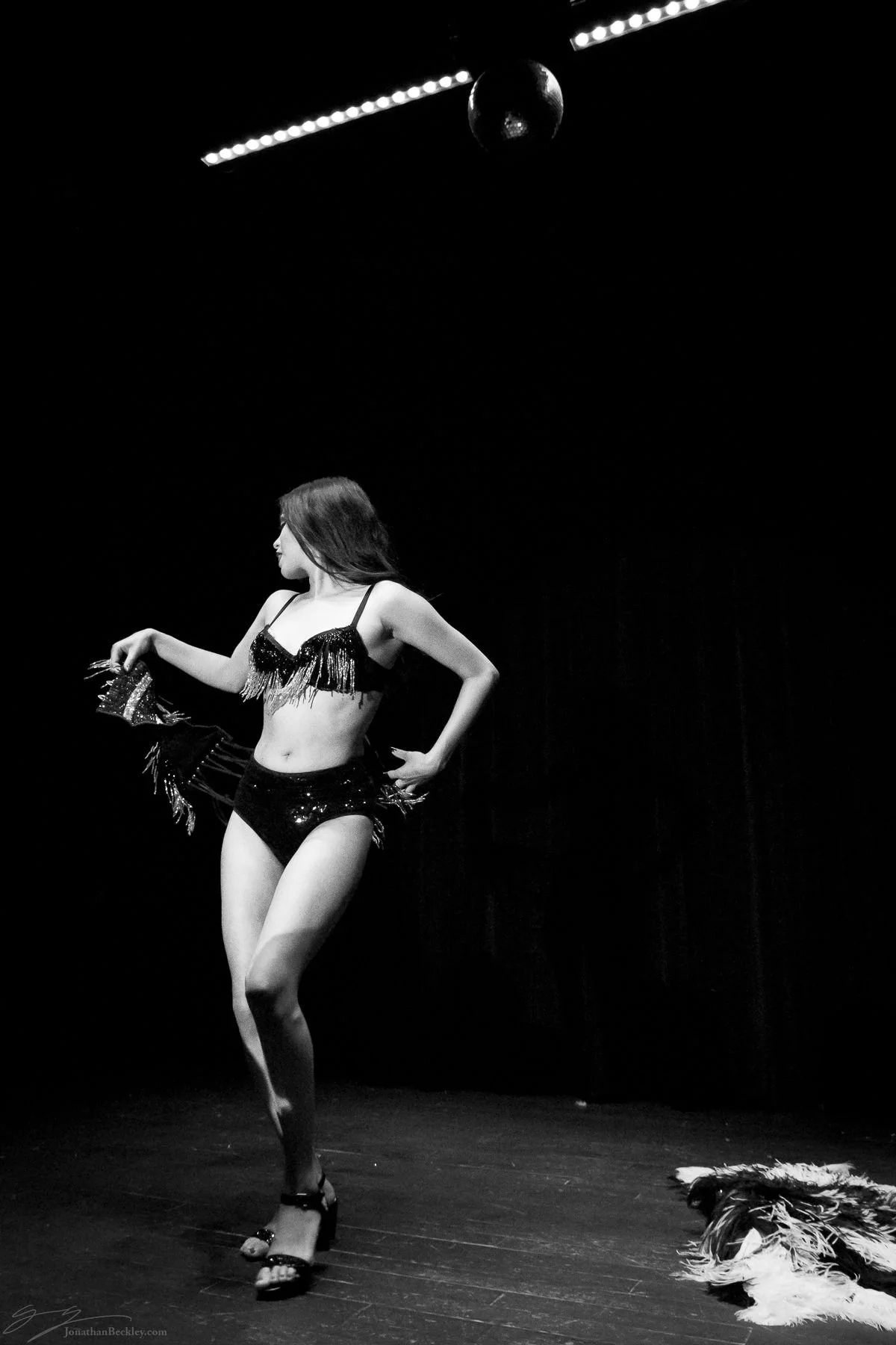 A woman on stage wearing a fringed, sequined dance costume, standing in profile, with a dark background and a disco ball overhead.