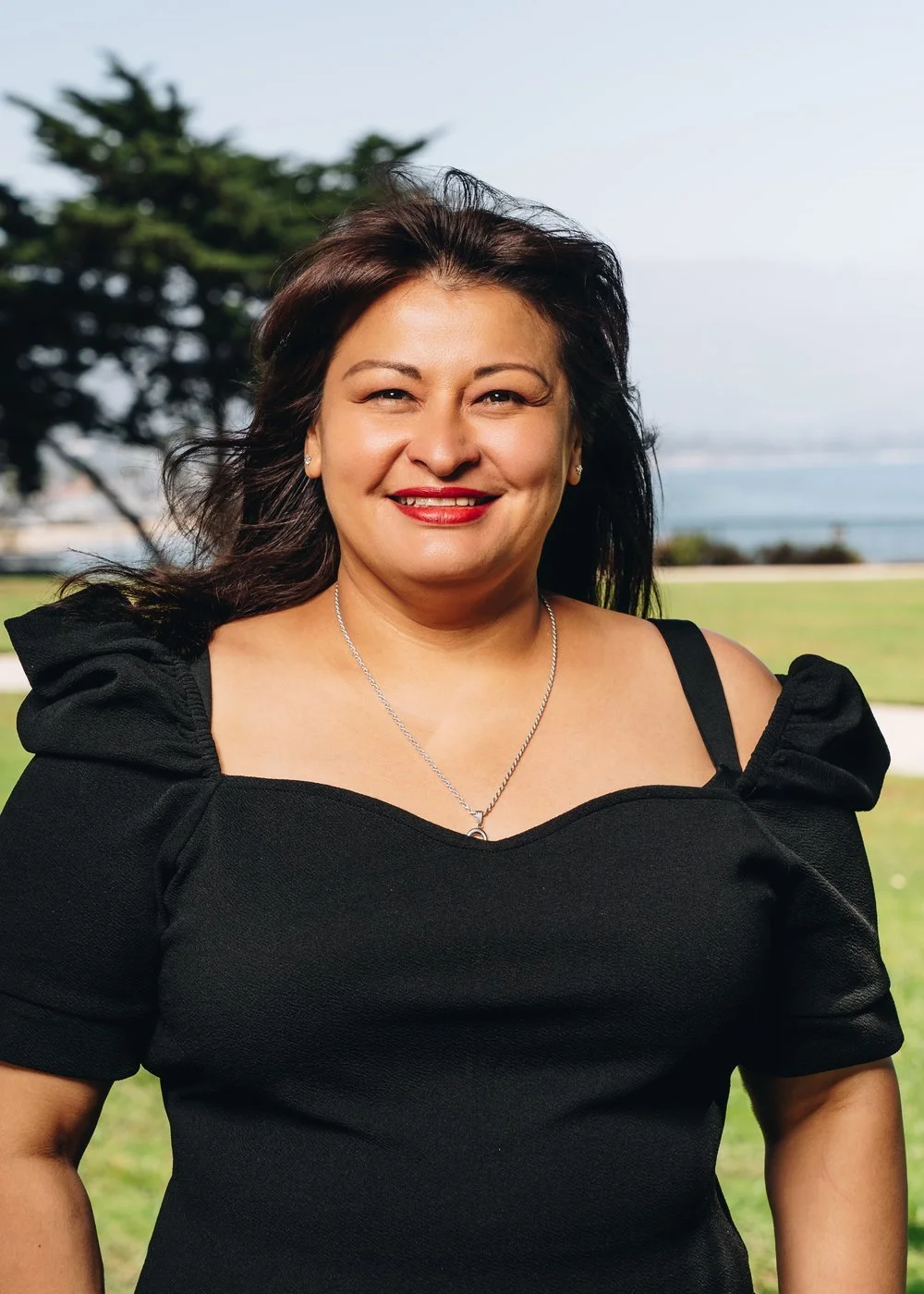 A smiling woman with dark hair in a black dress standing outdoors near a park with trees, grass, and water in the background.