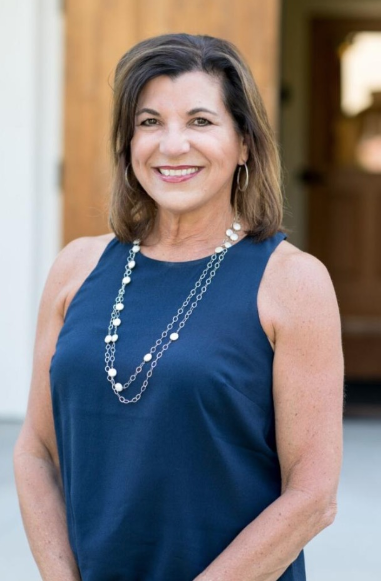 A smiling woman with shoulder-length brown hair, wearing a sleeveless blue dress and pearl jewelry, standing outdoors in front of a wooden background.