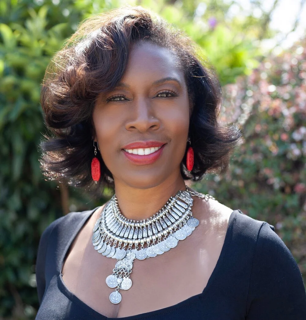 A smiling woman with dark hair styled in loose waves, wearing a black top, large red earrings, and multiple layered silver necklaces, standing outdoors with lush greenery and colorful flowers in the background.