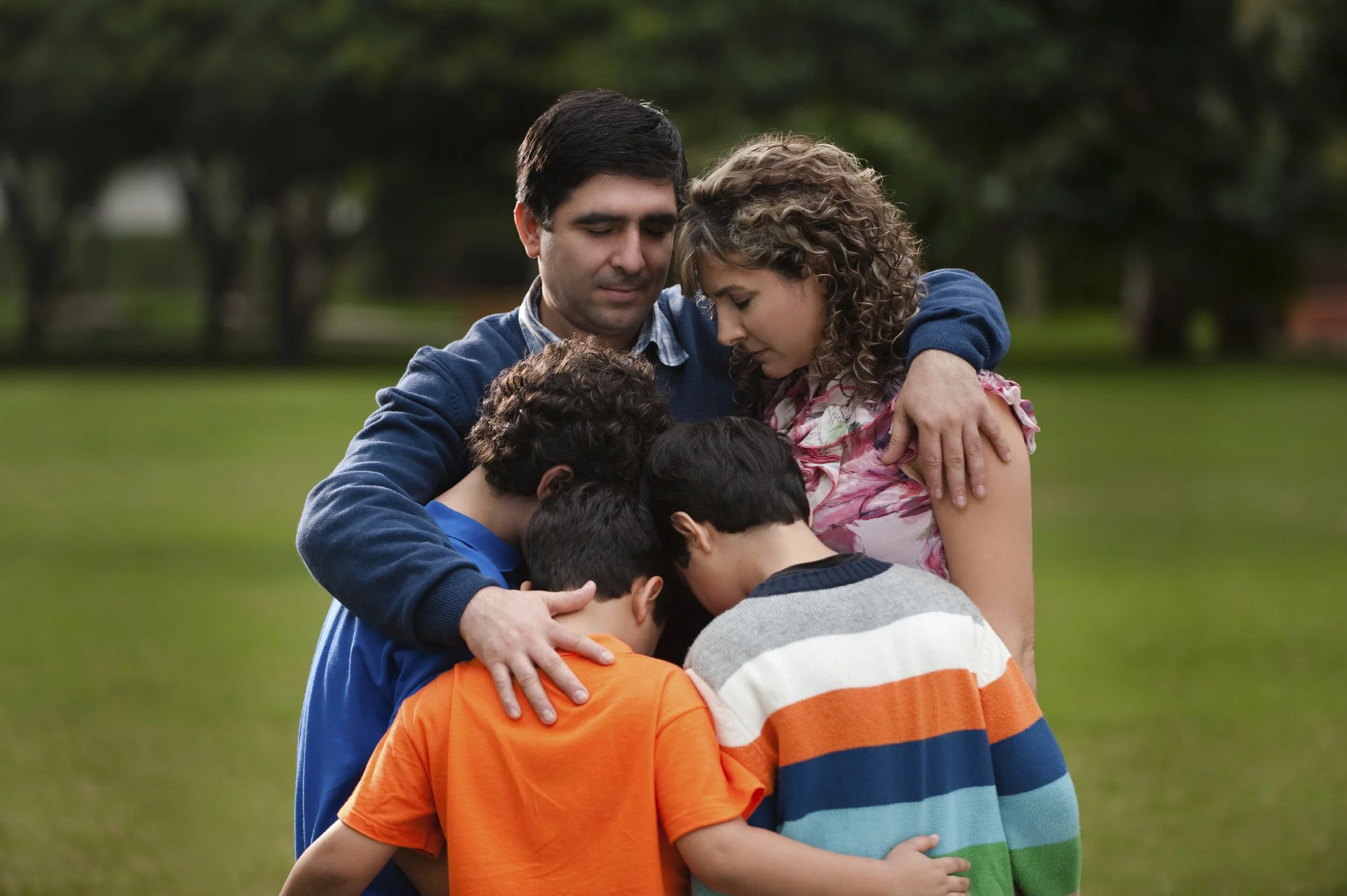 A family of six, including two adults and four children, standing outdoors on grass, with heads bowed together in prayer.