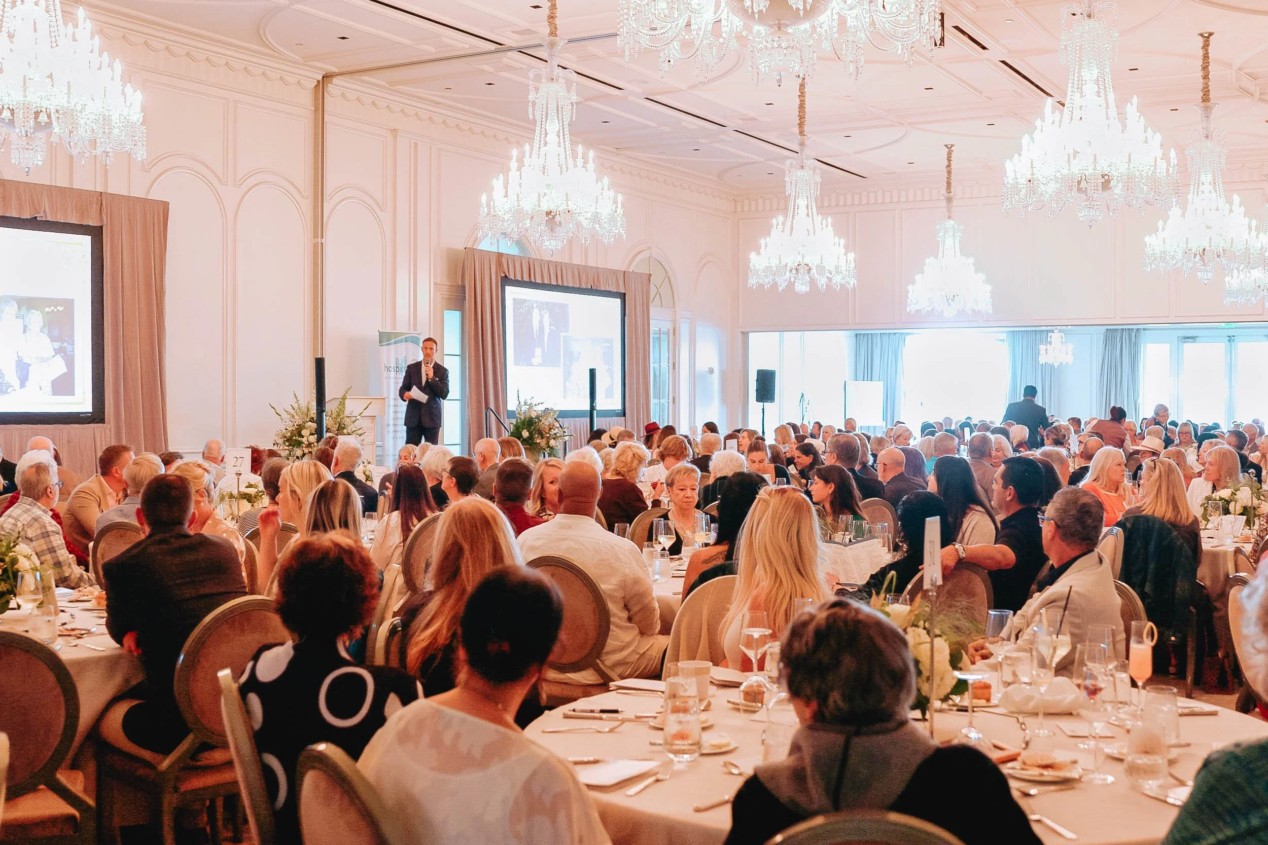 A large banquet hall filled with round tables and many attendees, with a presenter speaking on stage and large screens displaying slides, chandeliers hanging from the ceiling, and bright natural light coming through the windows.