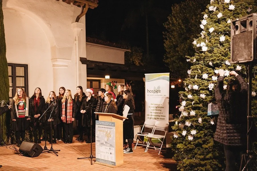 Group of people, including children, singing or performing at a Christmas event outdoors at night. A Christmas tree decorated with white ribbons and flowers, a banner for hospice of Santa Barbara, and a woman adjusting a speaker are visible.