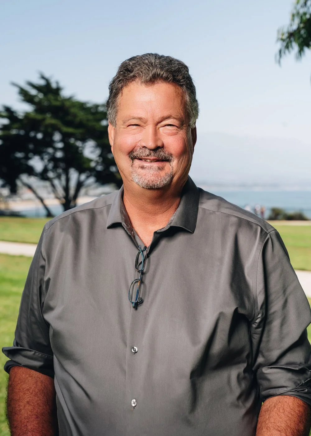 A smiling middle-aged man with gray hair and a beard, wearing a gray shirt and glasses hanging from the collar, standing outdoors with trees, grass, and a body of water in the background.