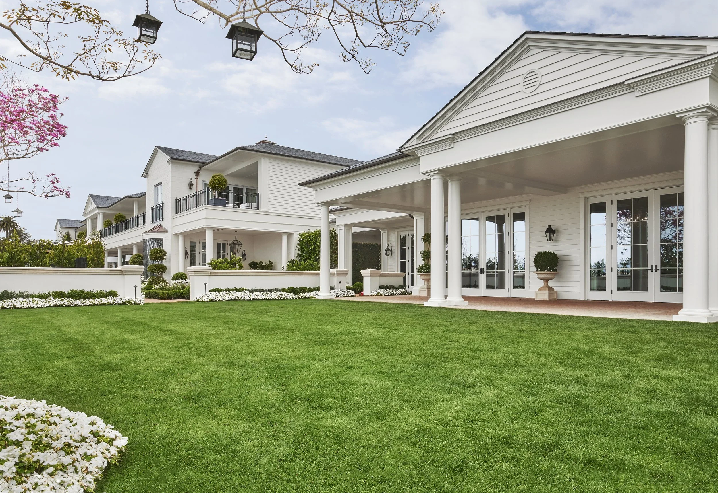 A large white house with a well-maintained lawn, decorated with potted plants, and surrounded by a white fence. The house has multiple balconies, large windows, and a covered patio with columns.