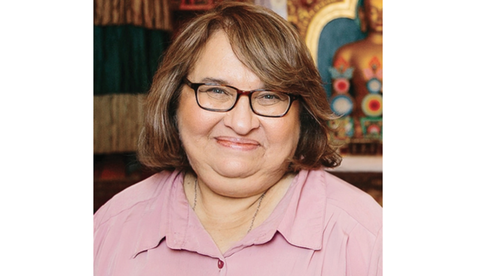 A woman with glasses and shoulder-length brown hair smiling, wearing a pink shirt, with colorful artwork in the background.