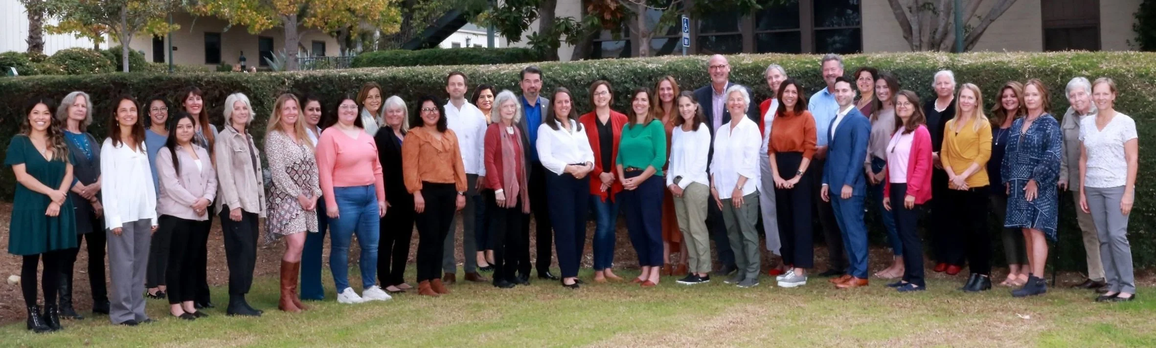 A large group of people standing outdoors on a lawn, posing for a photo in front of a hedge and trees during daytime.