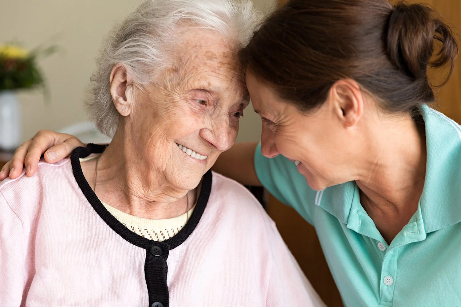 An elderly woman and a female caregiver lean their foreheads together, smiling and sharing a warm moment, with the caregiver's arm around the woman's shoulder.