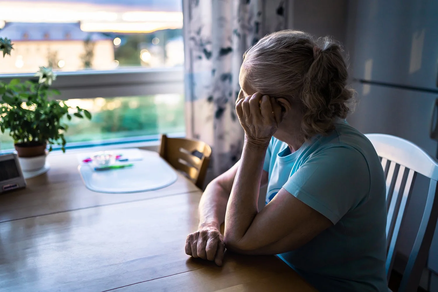 An elderly woman sitting at a dining table with her head in her hand, looking downcast, in a room with a window and a plant on the table.