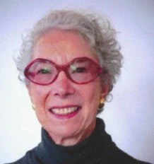 Portrait of an elderly woman with curly gray hair, red glasses, and gold earrings, smiling at the camera.