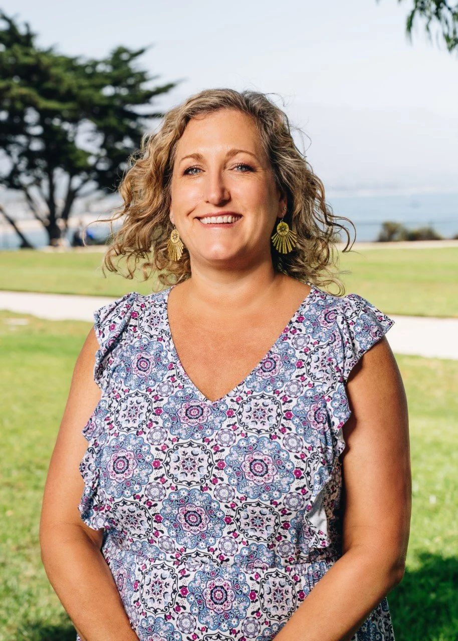 A smiling woman with curly blonde hair posing outdoors in a park near water, wearing a patterned sleeveless blouse and large yellow earrings.