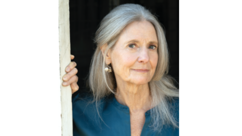 An older woman with long gray hair wearing a blue top, standing next to a wooden structure, with a dark background.