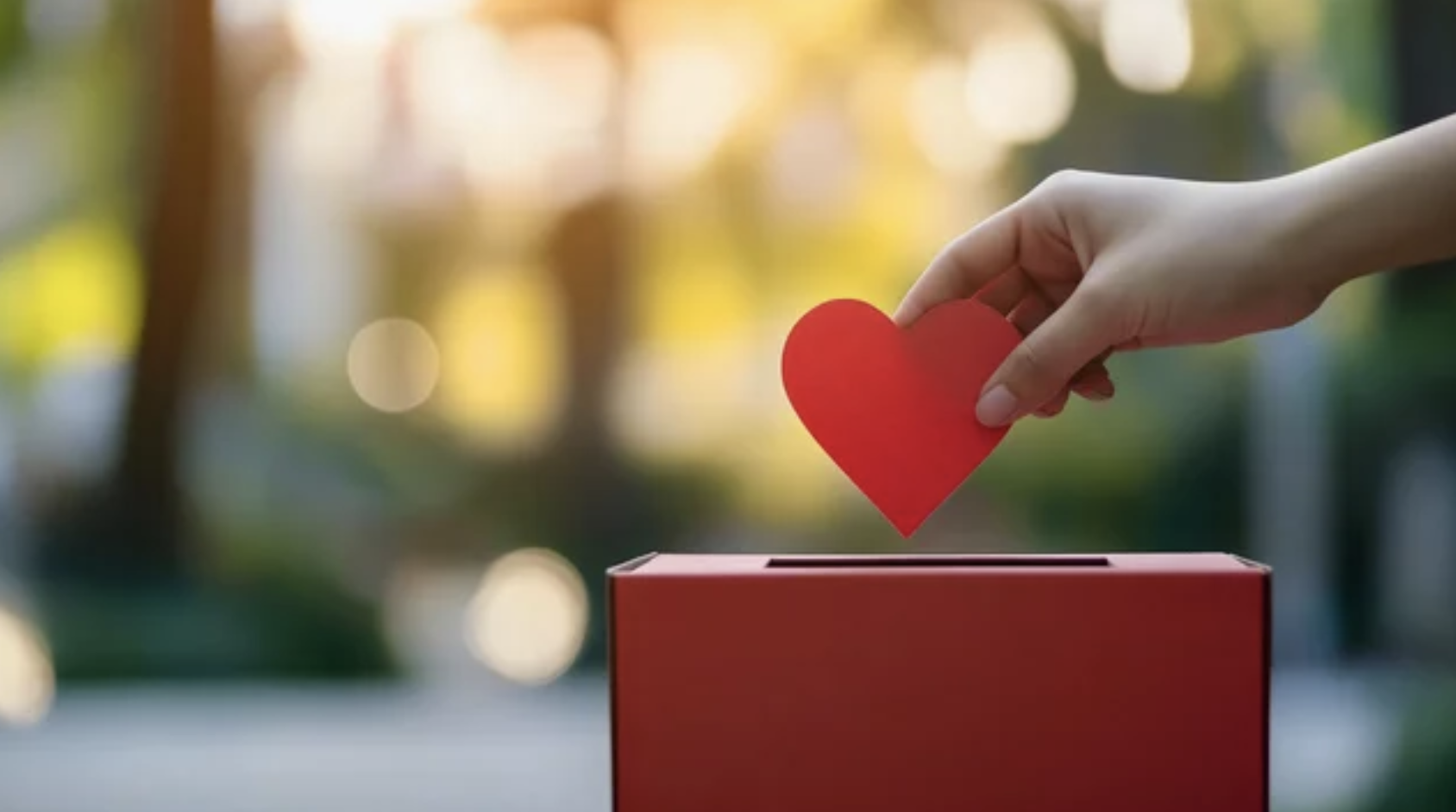 A hand placing a red paper heart into a donation box.