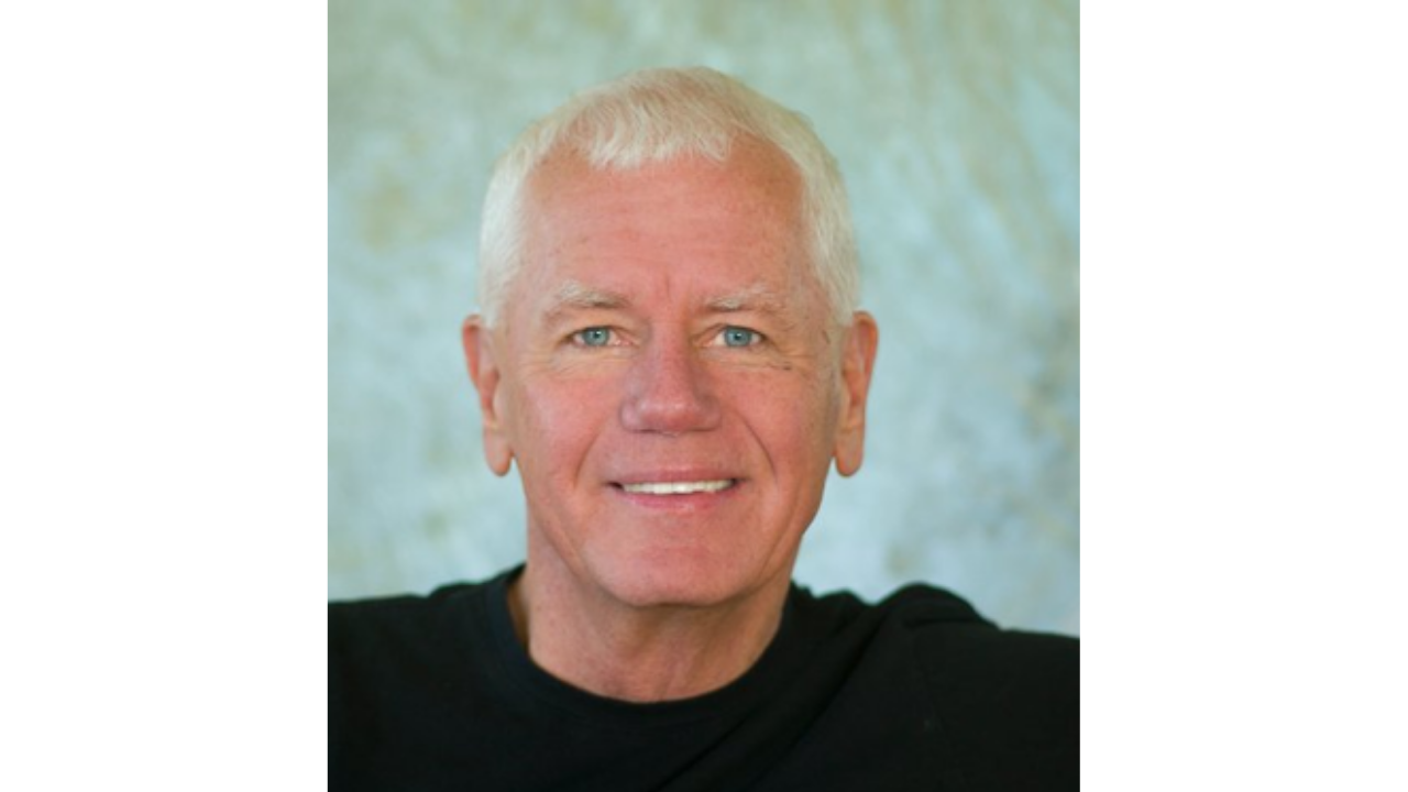Portrait of an elderly man with short white hair, blue eyes, wearing a black shirt, smiling, against a light, textured background.