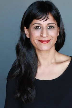 A woman with long black hair, wearing a black top, smiling with a neutral gray background.