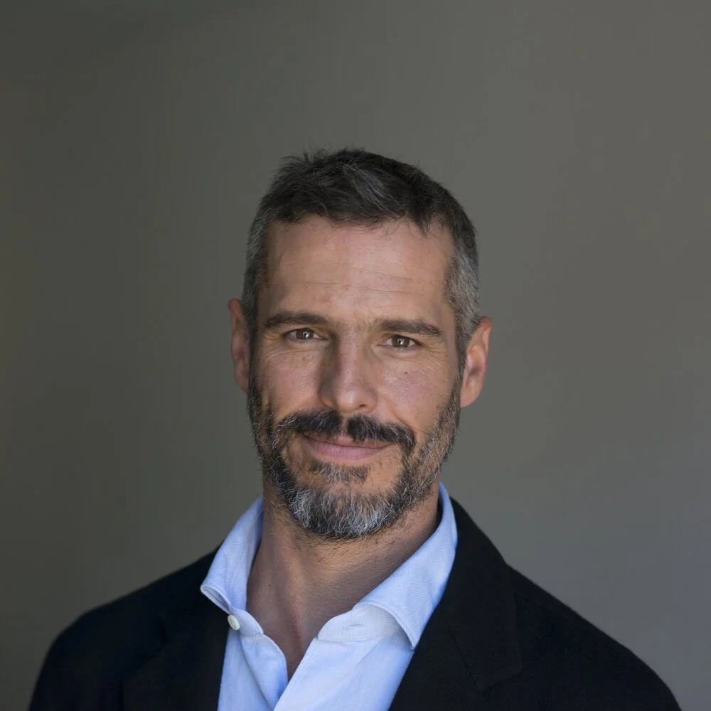 A middle-aged man with short, dark and gray hair, a beard, and blue eyes, wearing a light blue collared shirt and a dark blazer, smiling softly against a plain gray background.