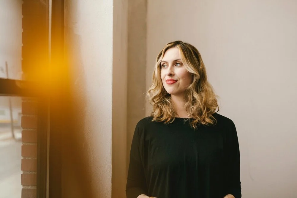 A woman with shoulder-length blonde curly hair, wearing a black top, standing indoors near a window with a brick column visible to her left.