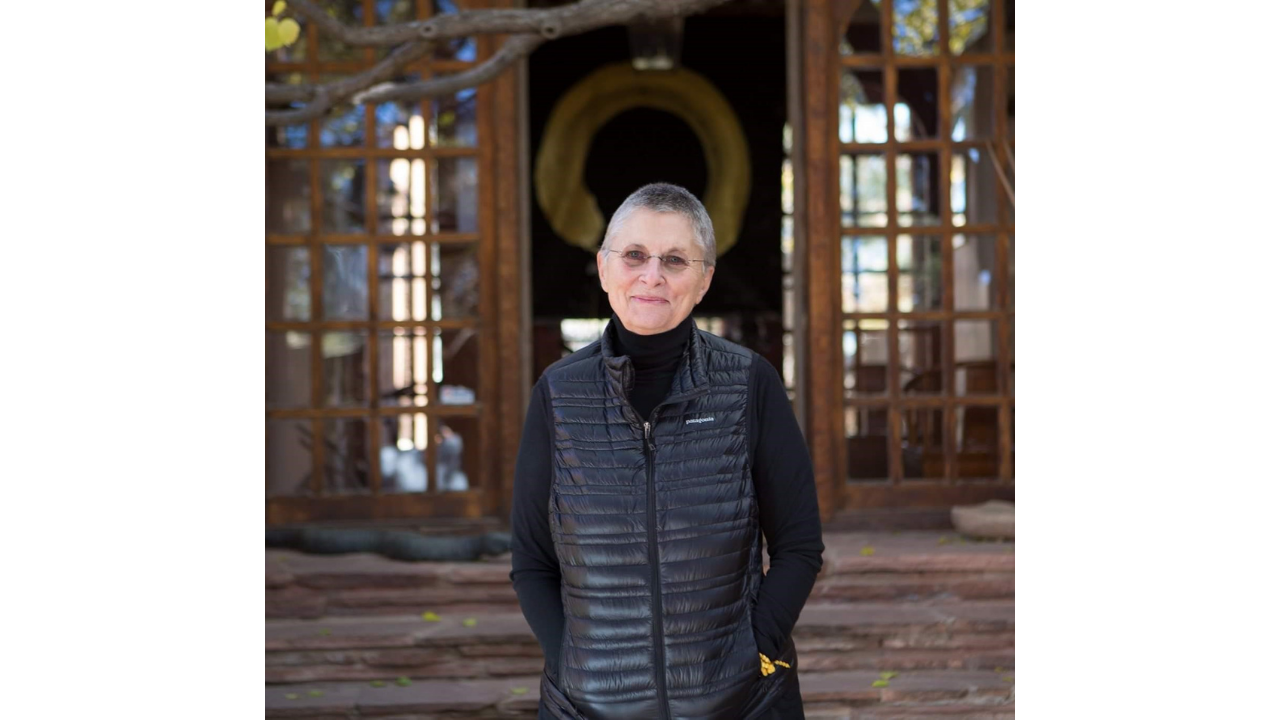 An older woman with short gray hair, glasses, and a black vest standing outside in front of a wooden structure with a circular opening.
