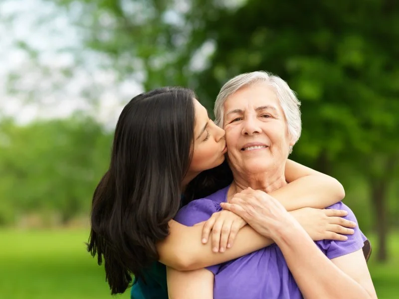 A young woman is kissing an older woman on the cheek outdoors in a park with green trees.