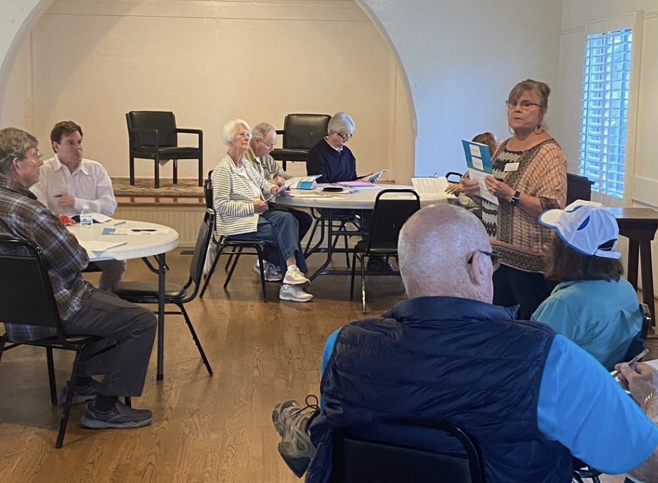 A group of elderly people sitting around tables and listening to a woman standing and speaking, in a room with hardwood floors and natural light.