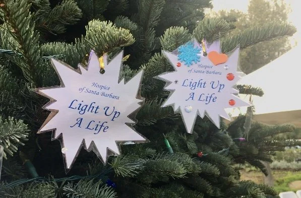 Two star-shaped ornaments hanging on a Christmas tree. The ornaments are white with a gold outline and have the text "Hospice of Santa Barbara Light Up A Life" written on them. One ornament has a blue snowflake, a pumpkin, and small red and white decorations attached.