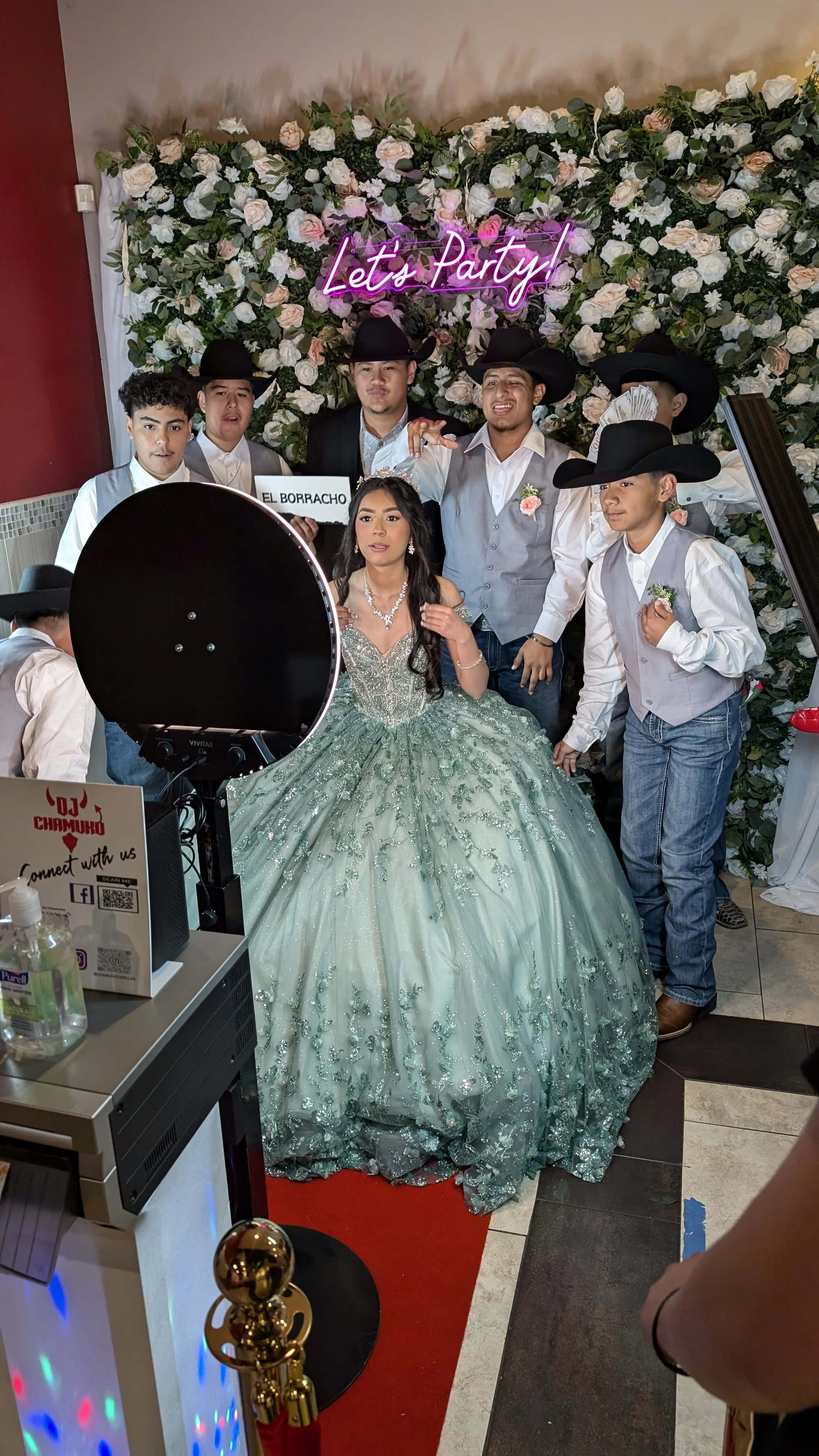 A group of people celebrating at a party with a floral backdrop and a neon sign that says 'Let's Party!'. A young woman in a green gown with intricate detailing is in the center, surrounded by boys dressed in cowboy hats and vests. There is a sign reading 'El Borracho' and a sign in the foreground that says 'DJ Chamuko'.
