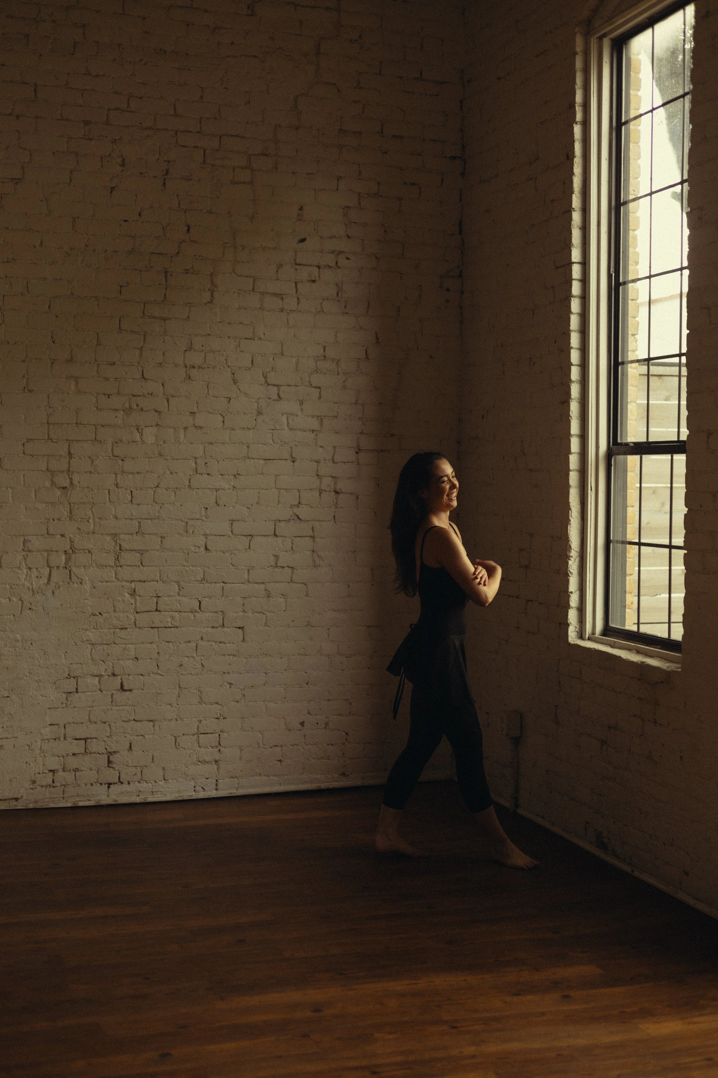 A woman in black clothing standing barefoot and smiling by a large window in a room with white brick walls and wooden floors.