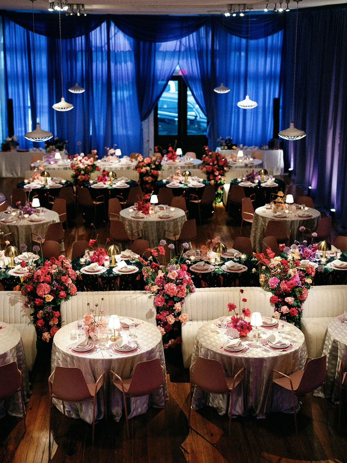 A decorated banquet hall with pink and purple floral arrangements, tables with pink napkins, and blue curtains.