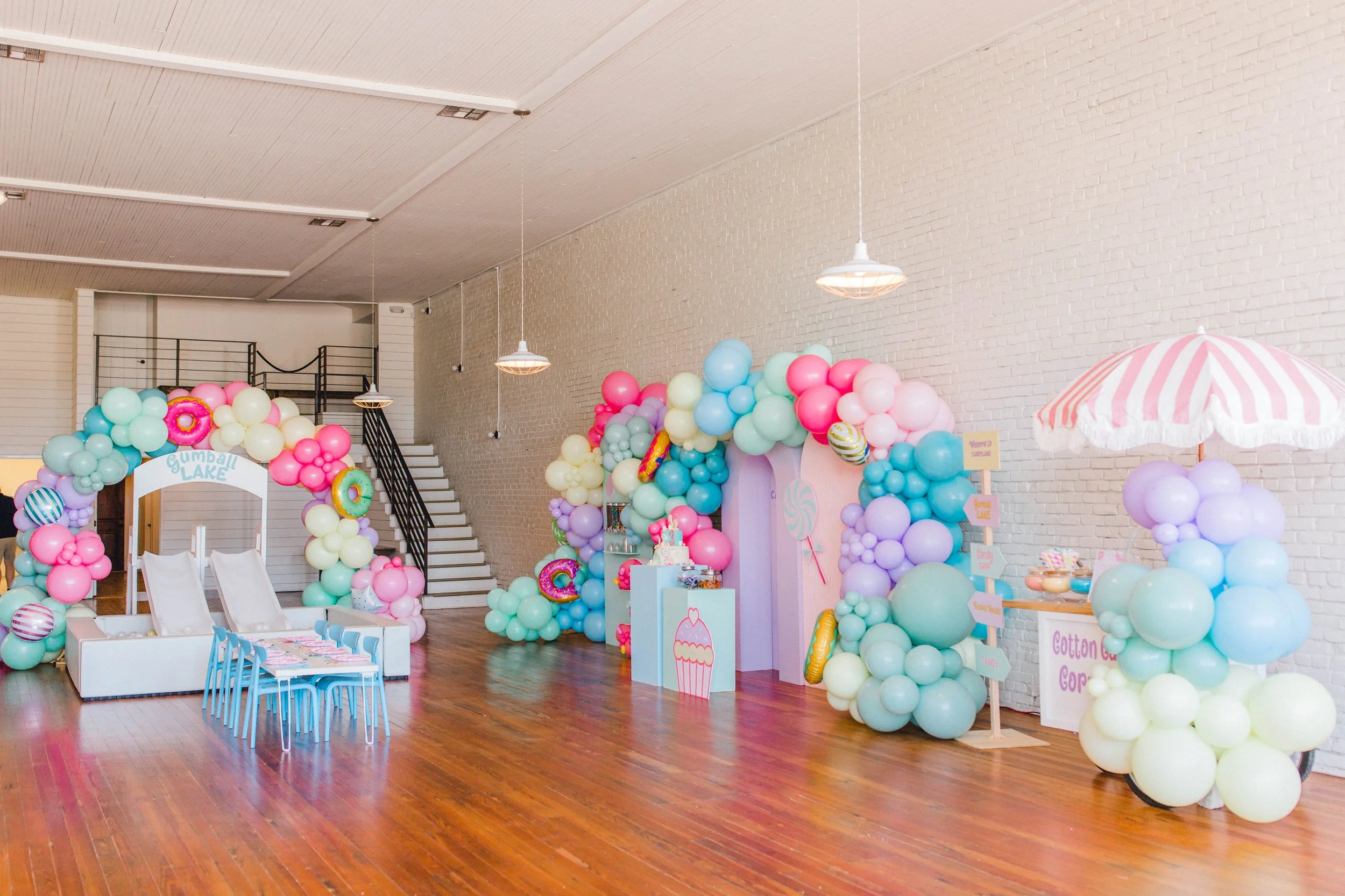 Decorated indoor space for a celebration with pastel-colored balloons forming an arch and various arrangements, including a small table with treats, a pink cupcake backdrop, and a cotton candy stand with a pink and white striped umbrella.