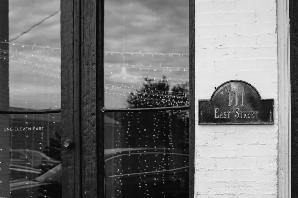 Black and white photo of a building entrance with a glass door, a street address plaque reading '111 East Street,' and the reflection of outdoor string lights and a tree in the glass.