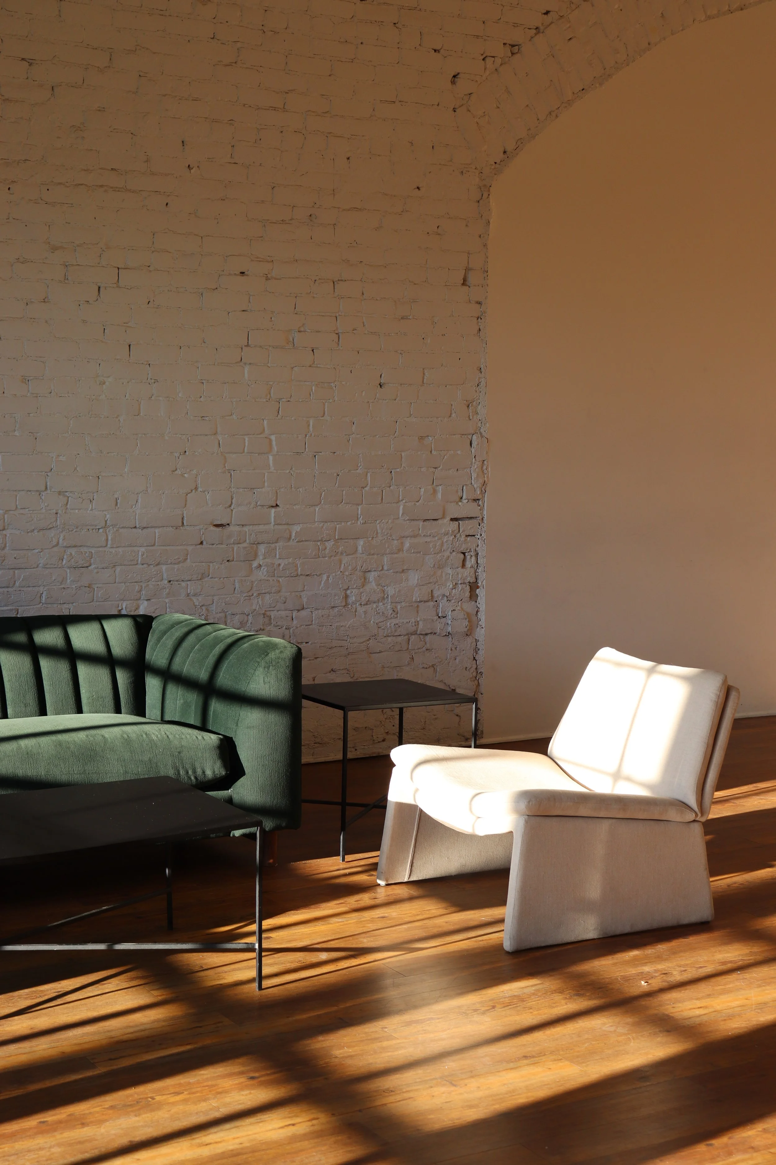 Sunlight streaming through window blinds cast shadows on a green velvet sofa, a white lounge chair, a black side table, and a black coffee table in a room with an exposed white brick wall and wooden floor.