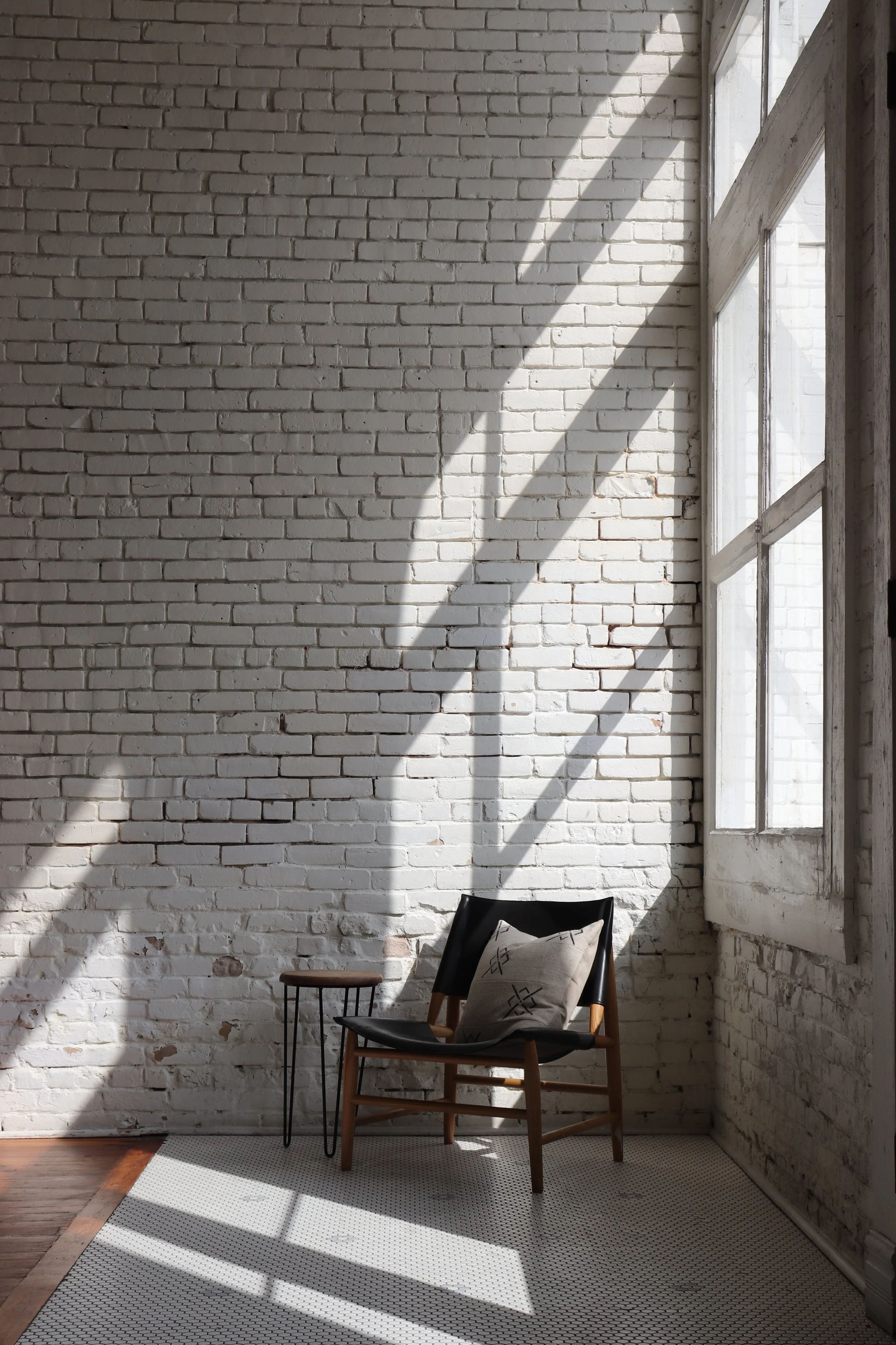 Sunlight streaming through large windows onto a white brick wall, casting shadows. A wooden chair with a black cushion and a decorative pillow is positioned on a small rug in the corner of a minimalistic room.