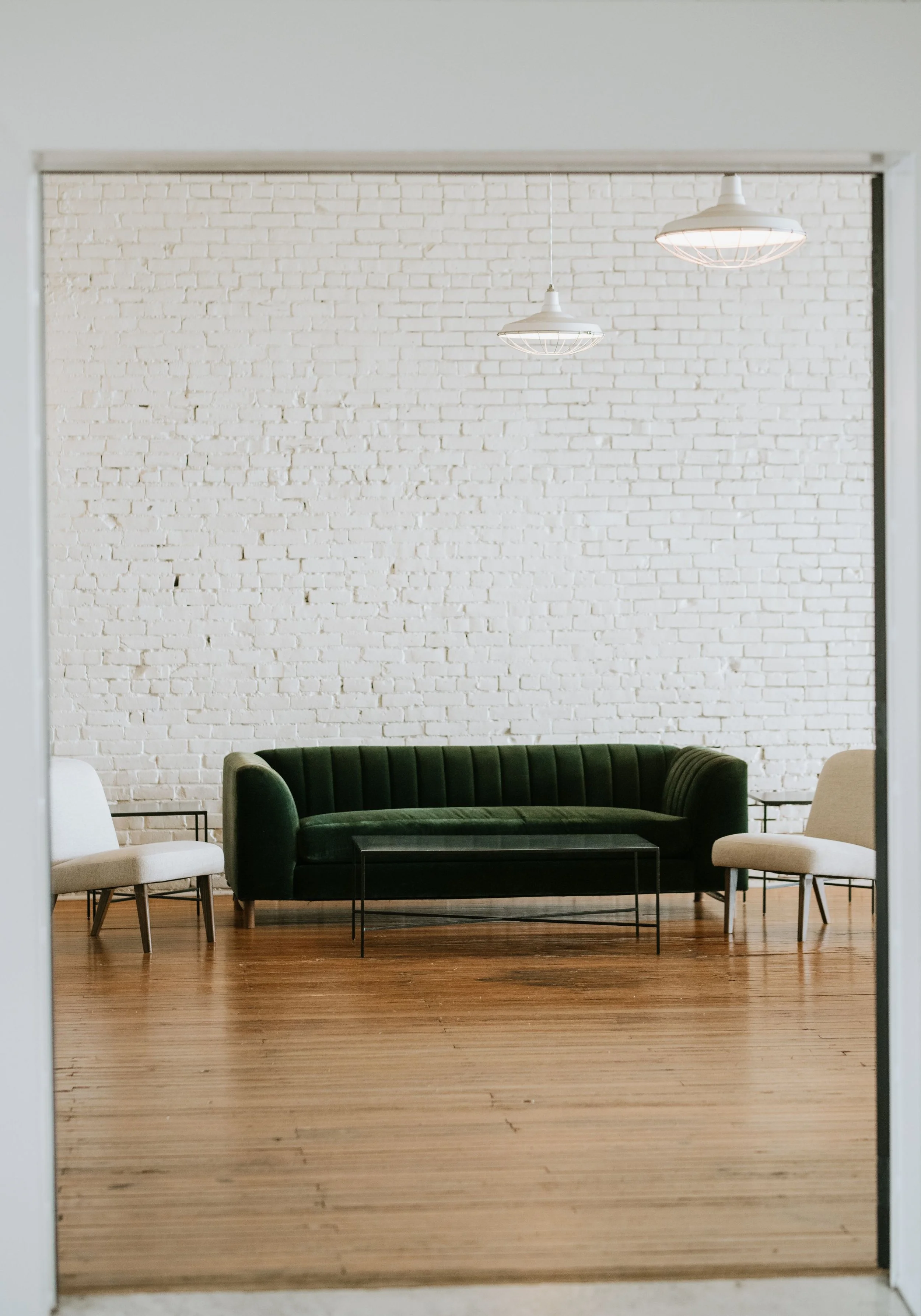 A minimalistic living room with a white brick wall, wooden floor, and three modern chairs in white, beige, and green. Two ceiling pendant lights hang from the ceiling.