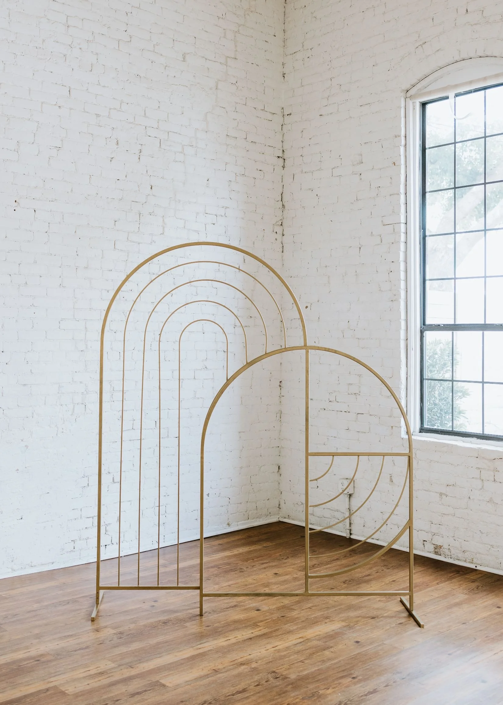 Decorative gold metal structure with geometric arches and curves standing in a bright room with white brick walls, wooden floor, and a large window.