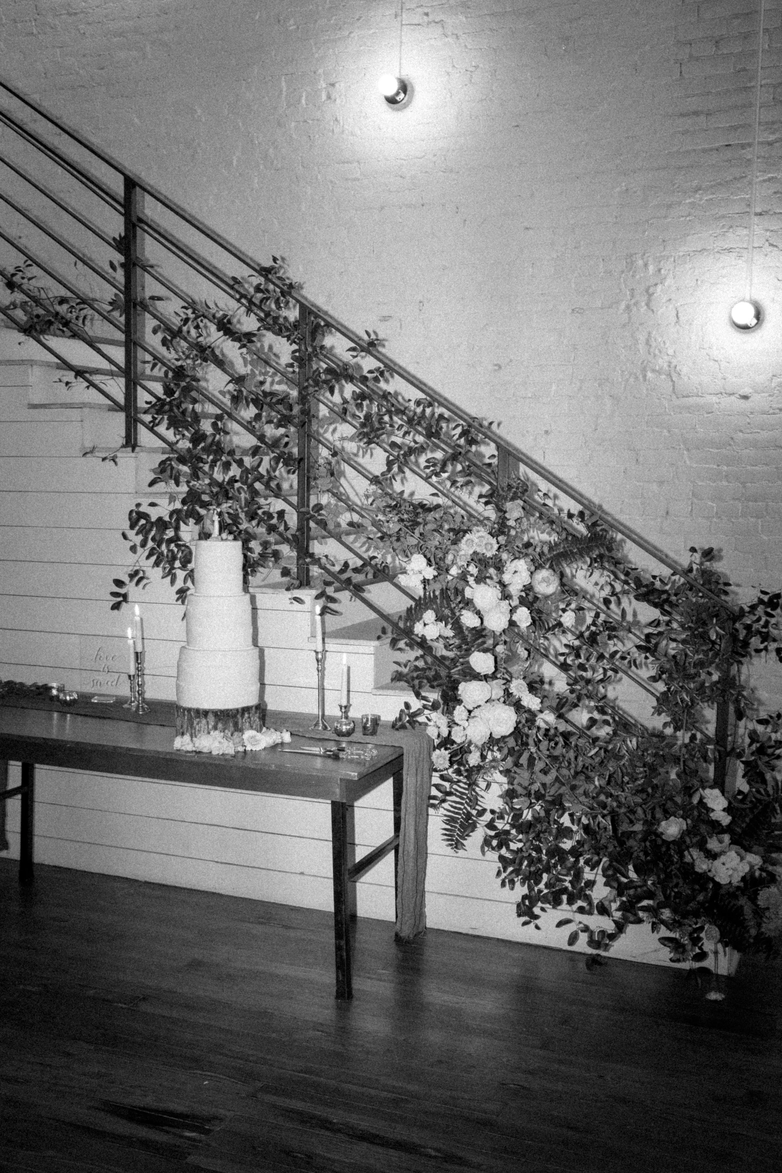 A wedding cake on a table with candles and flowers, set against a backdrop of a staircase decorated with greenery and flowers, in black and white.