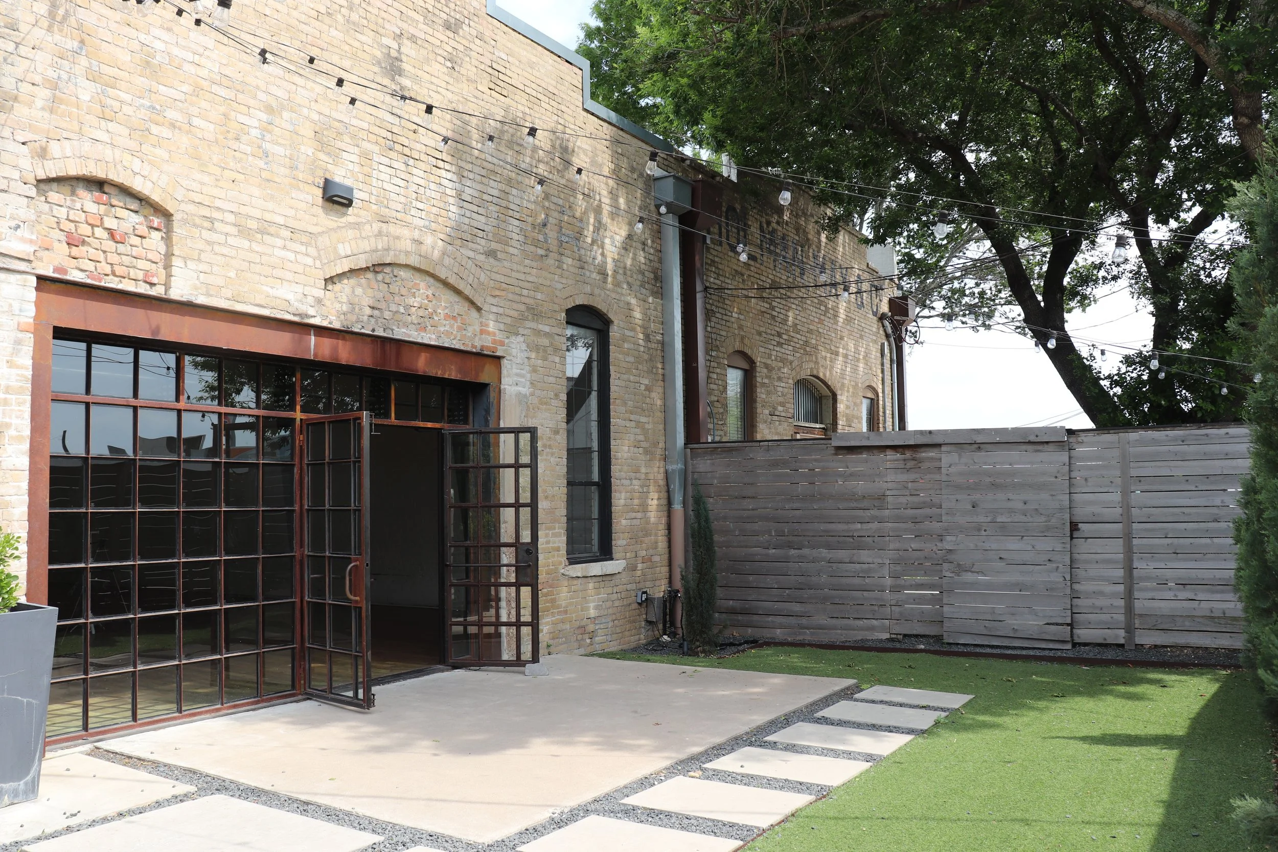 Backyard patio area with large glass doors, a wooden privacy fence, artificial grass, stepping stones, and string lights, adjacent to an old brick building.