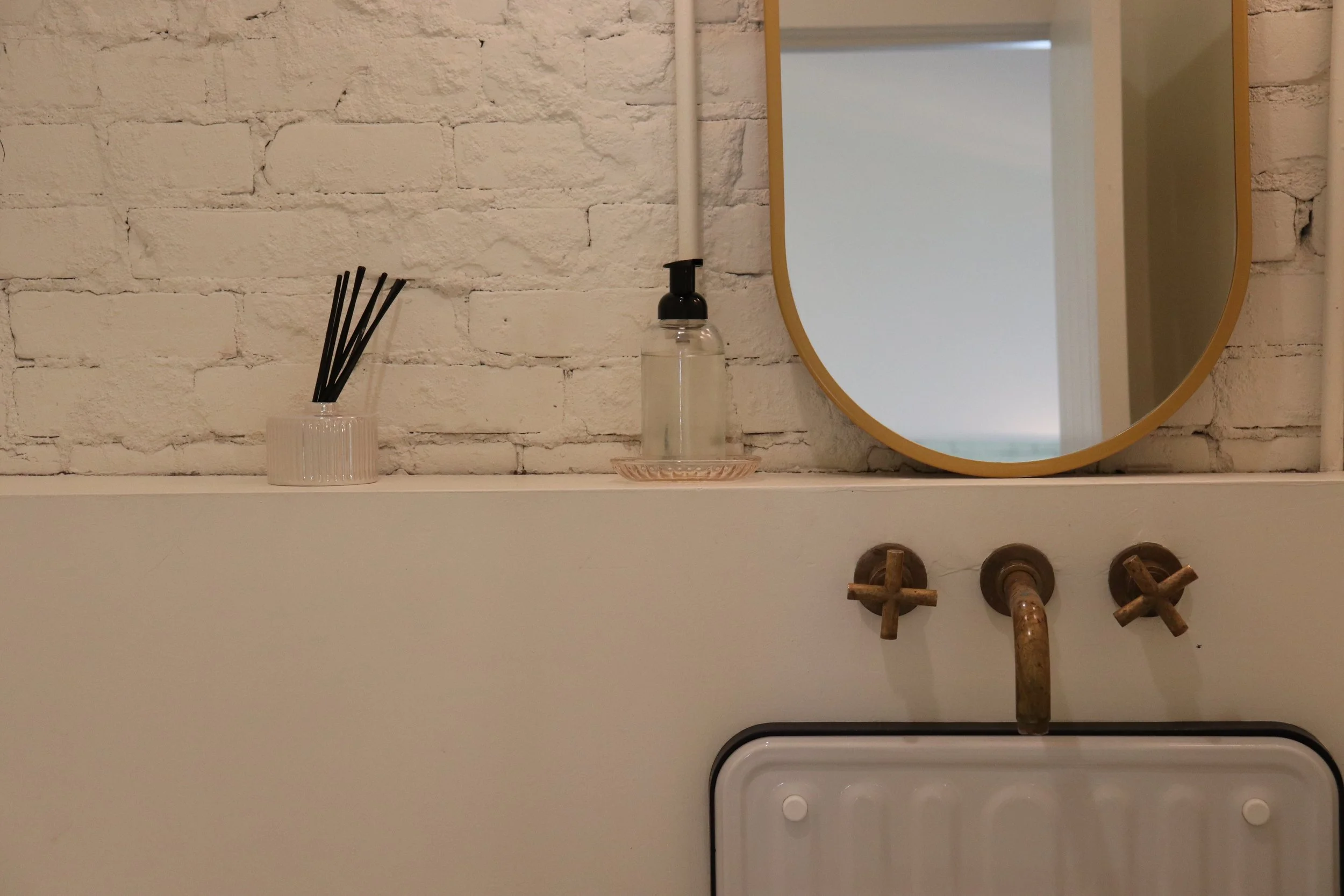 A white shelf with a small white container holding black reed diffusers, a transparent soap dispenser with a black pump, and a round mirror with a gold border against a white brick wall.