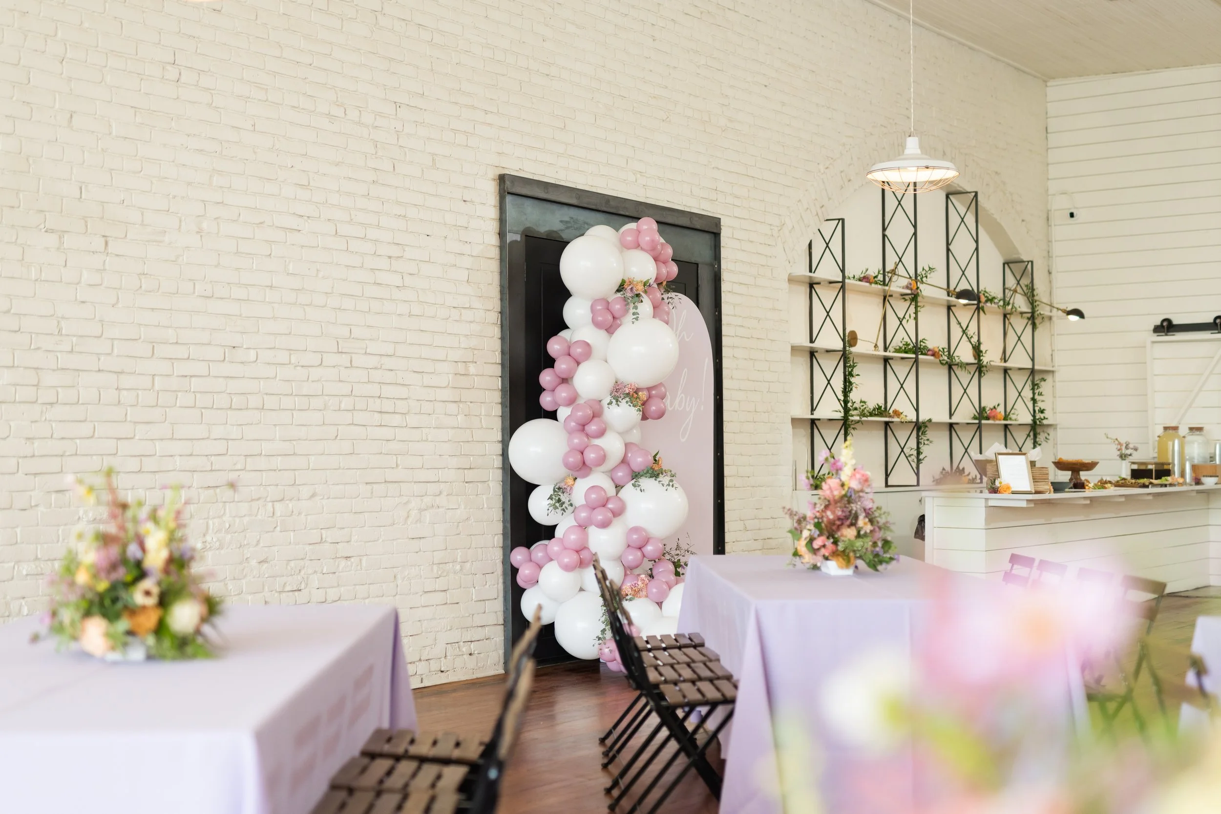 Decorated event space with pink, white, and purple balloons forming an arch near a sign, pink tablecloths with floral centerpieces, wooden chairs, and a buffet area against white brick walls.