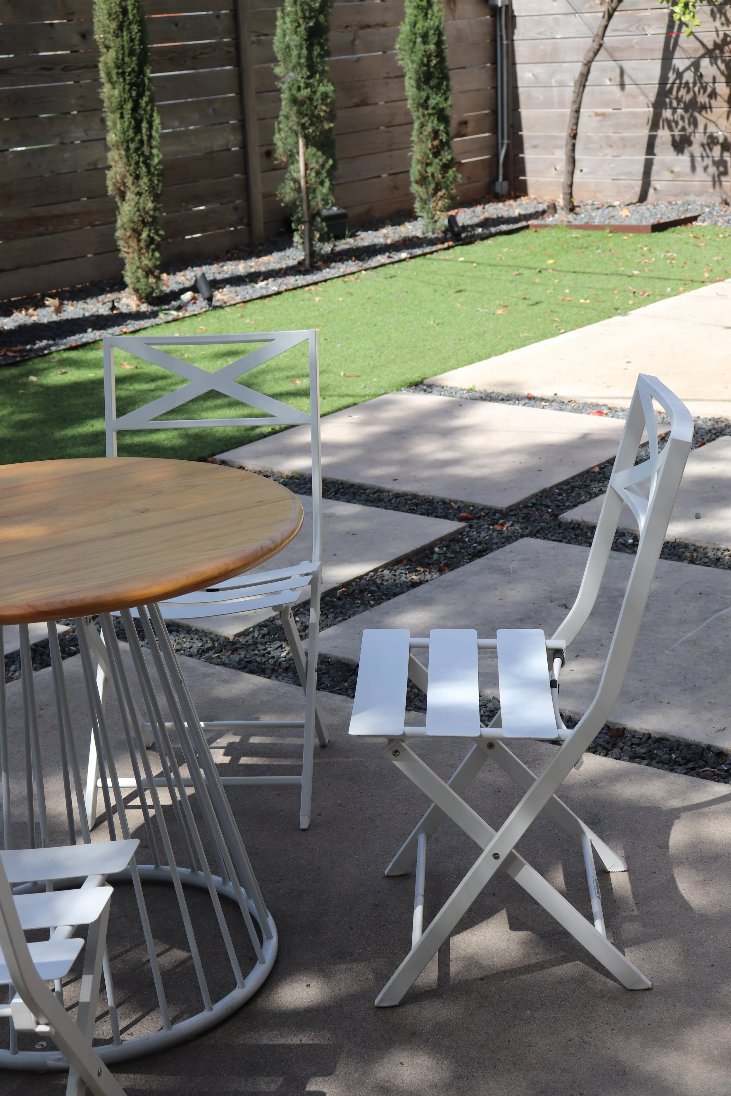 Outdoor patio with a round wooden table and white folding chairs, set on a concrete surface, with a small grassy area and a wooden fence in the background.